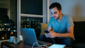 A man looks exhausted and overwhelmed while sitting on a couch at night, surrounded by a laptop, a smartphone, and a tablet.