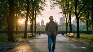 A smiling middle-aged man walks along a wet, tree-lined path in an urban park, likely Central Park, during the golden light of sunrise with a city skyline visible in the background.